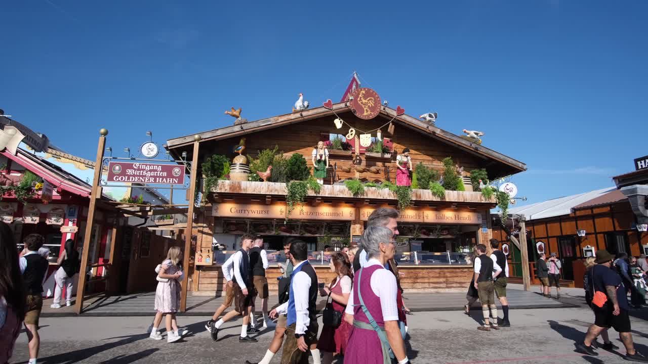 An establishment inside Theresienwiese, adorned with a traditional German statue, stands proudly as people gather in front, admiring Oktoberfest's detailed decorations and lively atmosphere.