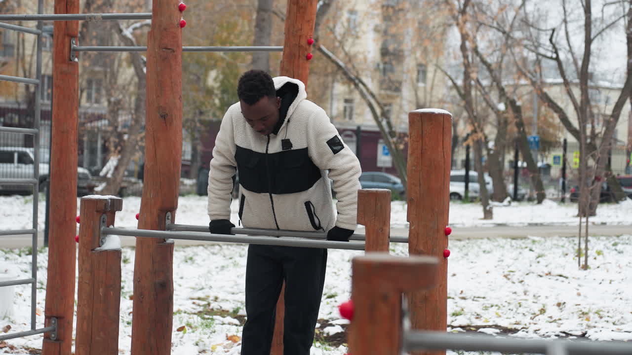 흑인 남성은 겨울 동안 바에서 운동 장비를 사용하여 피트니스 루틴에 참여합니다. 눈이 많은 바쁜 도시 배경 속에서 결단력과 도시 생활 방식을 보여줍니다.