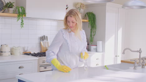 panning shot of woman at home in kitchen wearing rubber gloves cleaning down work surface using cleaning spray - shot in slow motion