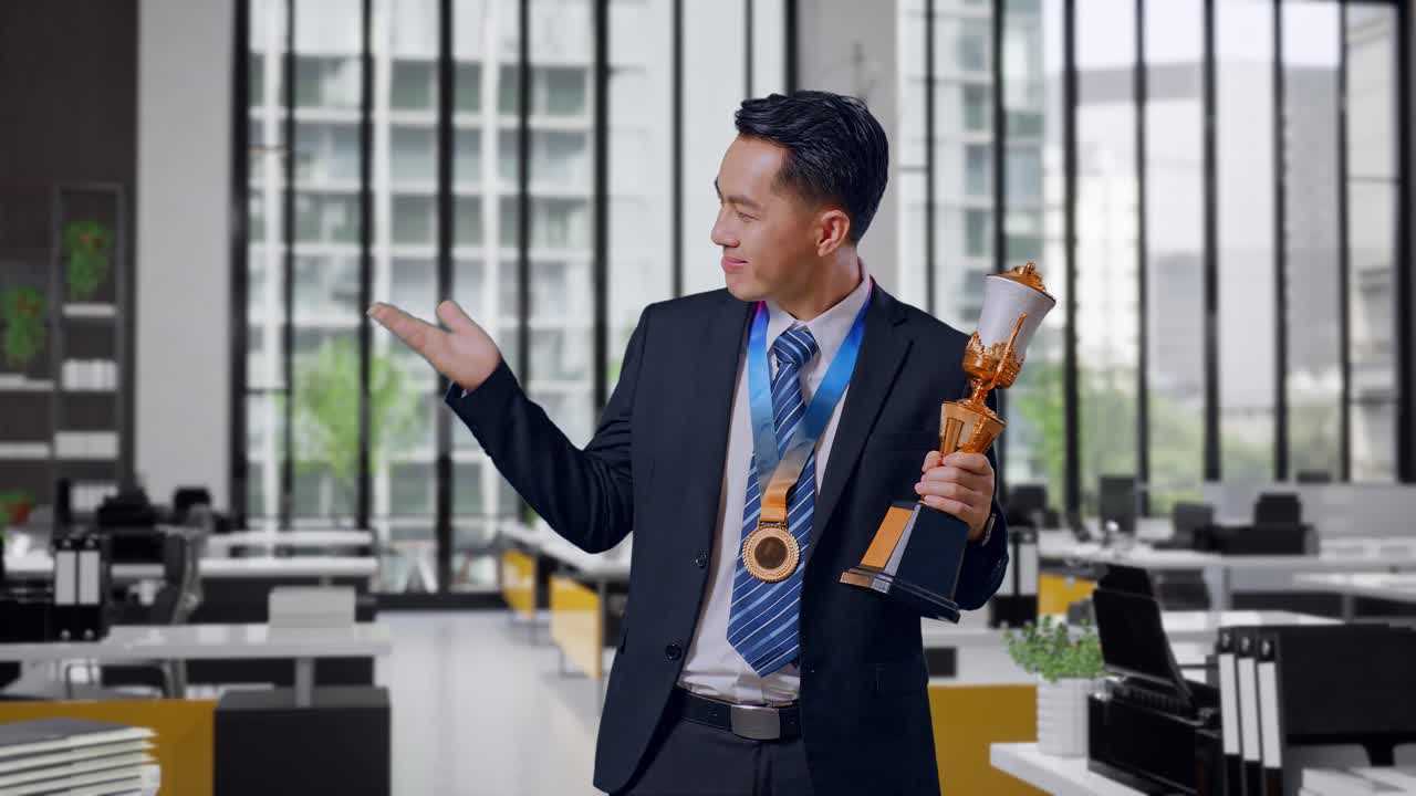 Asian Business Man In A Suit And Tie With A Gold Medal Looking At A Gold Trophy In His Hands, Smiling, And Pointing To Side In Office