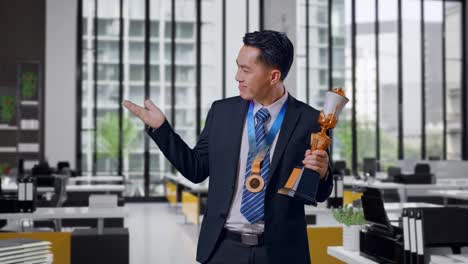 asian business man in a suit and tie with a gold medal looking at a gold trophy in his hands, smiling, and pointing to side in office