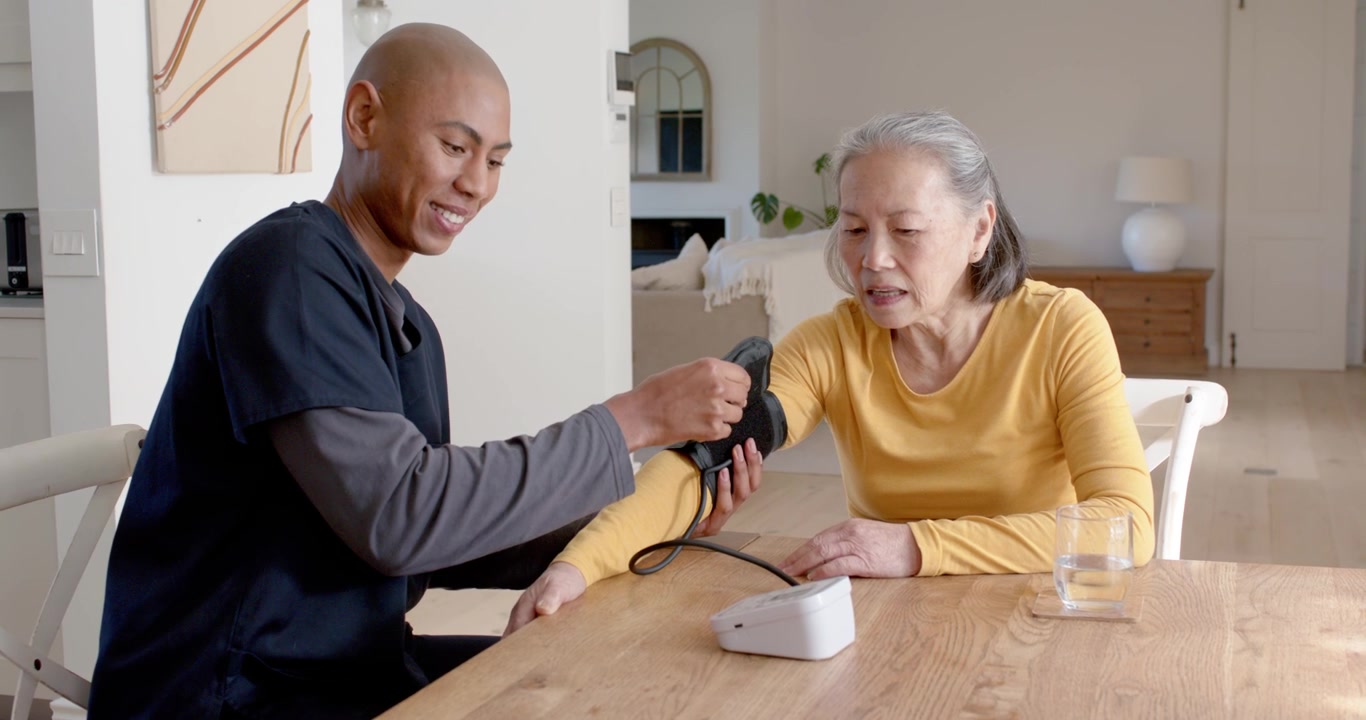 Donna asiatica anziana e caregiver in divisa da lavoro che applica un bracciale al tavolo con monitor per la misurazione della pressione sanguigna