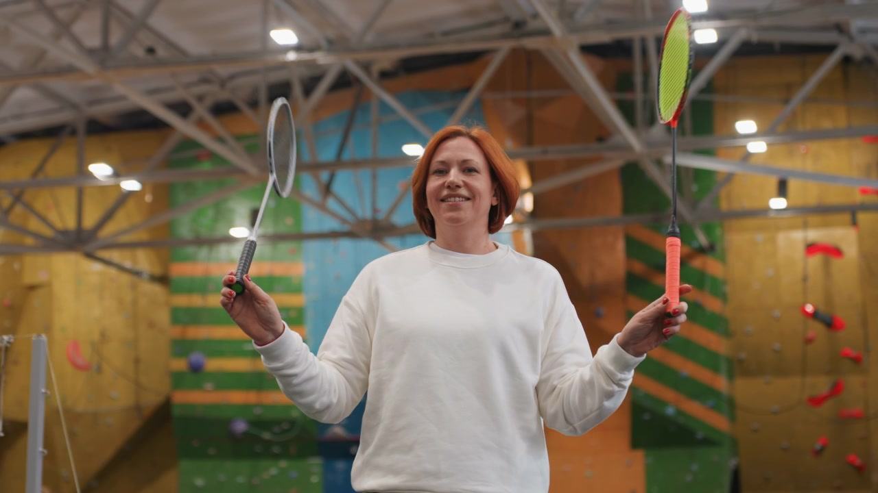 Smiling woman juggling rackets. Energetic female athlete playing sports happily. Cheerful female player holding two rackets in vibrant climbing gym atmosphere with bright illumination