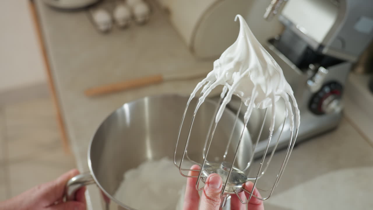 Close up of whisk mixer coated with thick whipped flour mixture held above mixing bowl, highlighting smooth texture and stiff peaks, perfect for baking, pastry prep, or dessert making in kitchen