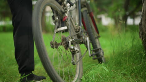 close-up leg view of individual in black trousers and sneakers walking up to parked bicycle in grassy field, lifting back tires and pedaling, causing tires to rotate briefly before placing it down