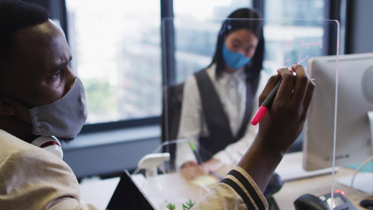 African american man wearing face mask writing on glass board at modern office