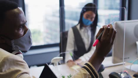 African-american-man-wearing-face-mask-writing-on-glass-board-at-modern-office