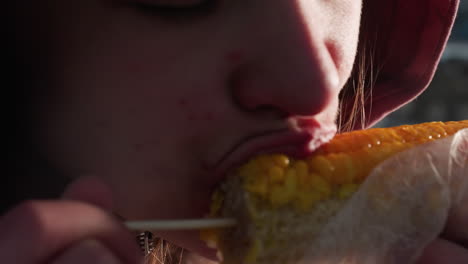 close-up of woman eating corn held by stick wrapped in white plastic with bokeh lights in blurred background, steam rising from freshly cooked corn in outdoor setting on sunny day