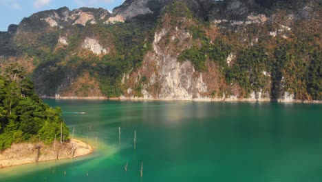 Viridian-Water-Of-Cheow-Lan-Dam-With-A-Beautiful-Mountains-On-The-Background-And-Two-Longtail-Boats-Sailing-On-The-Lake-In-The-Distance,-Thailand