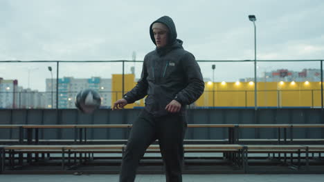 athlete juggling ball with knee during training in urban sport arena, demonstrating focus, against a modern cityscape with benches, buildings, and outdoor fitness equipment in the background