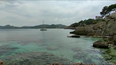 Küste-Auf-Palma-De-Mallorca-In-Der-Nähe-Von-Palma-Cape-De-Pera,-Chistmafrys-Mit-Einem-Segelboot-Auf-Dem-Wasser,-Ein-Haus-Auf-Einem-Berg-Bei-Gutem-Wetter