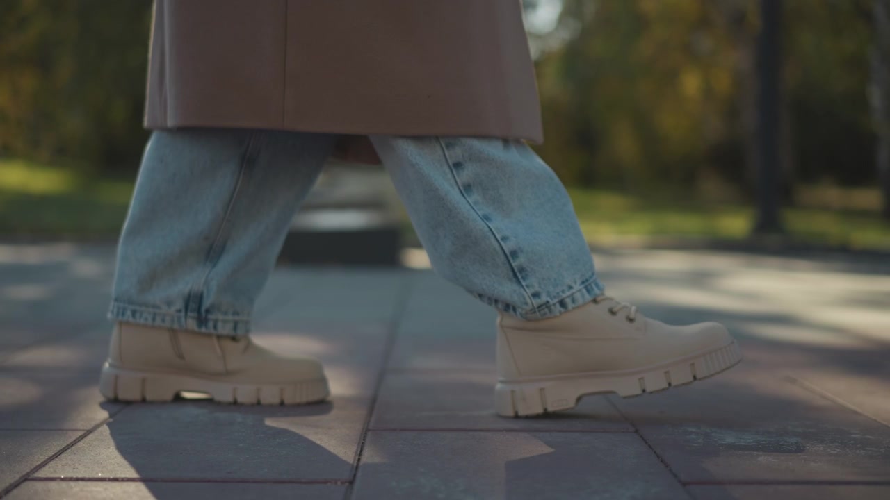 Close up of person feet in light blue jeans and beige boots walking on paved ground with soft sunlight casting shadows. subject wears rolled up denim pants revealing lower legs as they take