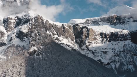 una vista aérea del cirque du fer à cheval mientras está cubierto de nieve durante un frío invierno, inclinándose hacia los picos nublados del circo, revelando las cascadas de nieve que gotean por los altos acantilados