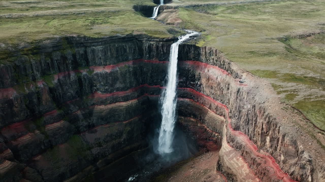 Wide aerial drone shot of Hengifoss Waterfall in motion, Iceland, sunny day, green grass, red clay. Part 2.