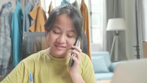 Close Up Of Asian Young Girl Online Seller Talk On Mobile Phone And Take Note While Using Computer For Selling Clothes At Home