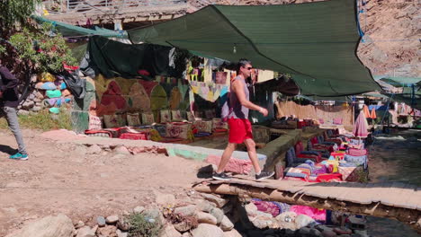 two men walk through an outdoor market in ourika under sunny skies with colorful stalls around them