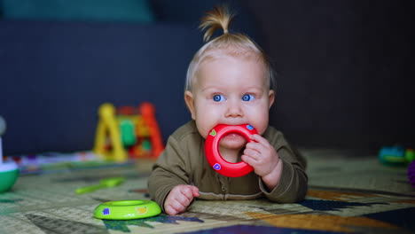 funny cute baby boy with blond hair lies on the floor. lovely infant chews a red toy ring as his teeth grow.