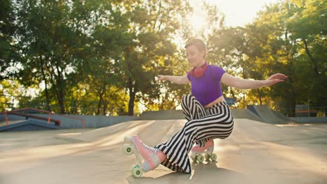 a girl with a short haircut in a purple top and striped pants rides sitting on roller skates in a skate park in summer. roller skating tricks at the skate park