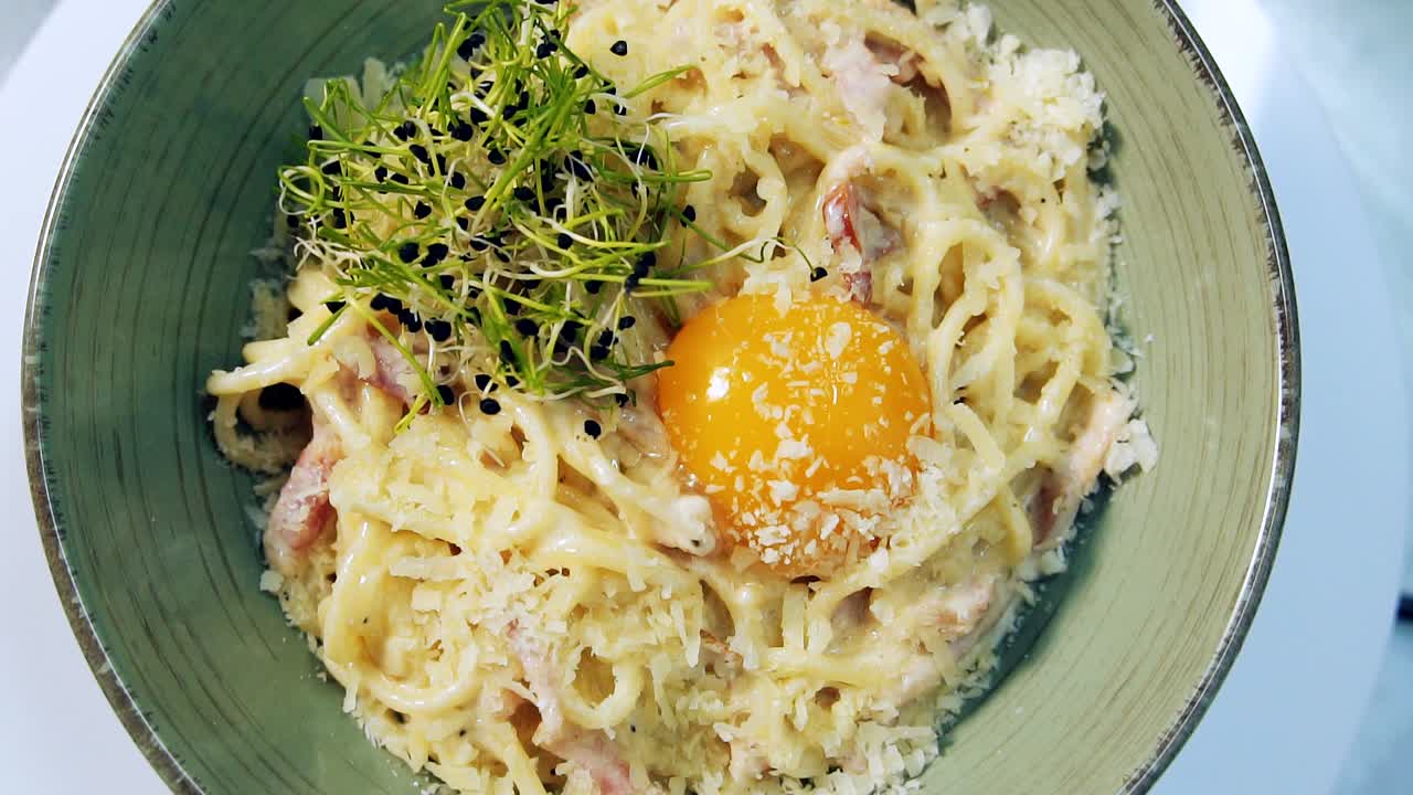 Rotating plate of a ready fresh-made pasta. Delicious spaghetti carbonara with yolk and cheese on top in a plate spinning. Yummy dish decorated with green sprouts. Close-up