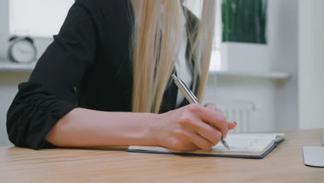 female writing with pen in notepad. crop view of woman with long blond hair in bright casual shirt sitting at wooden desk and writing down information with shiny metal pen into daily planner at day.