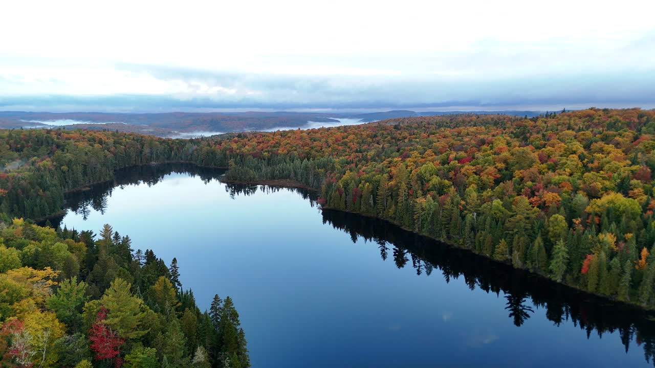 Drone view of a colorful autumn forest with mountains, lake, and river at sunrise in Mauricie, Quebec, Canada. Warm morning light highlights vibrant fall foliage and peaceful landscape