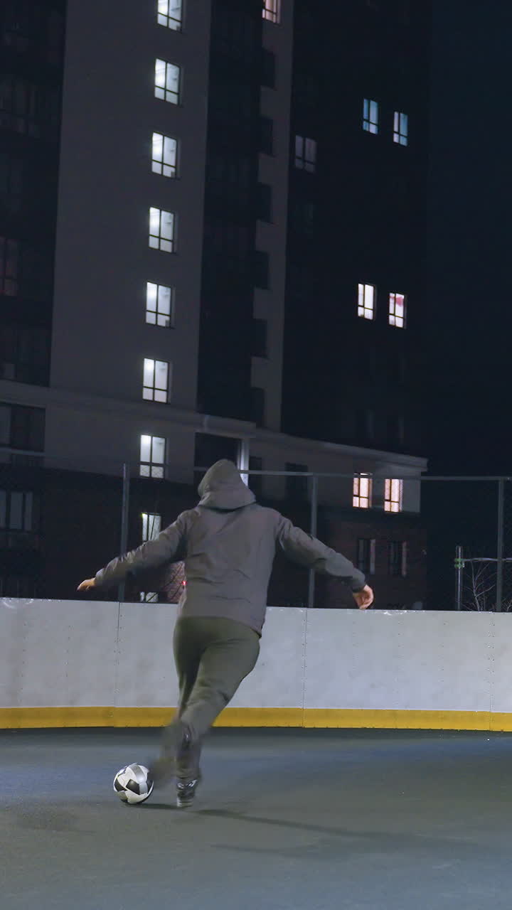 Man kicks soccer ball toward goalpost under artificial night lighting on outdoor court, with urban background and partial view of person walking behind fence
