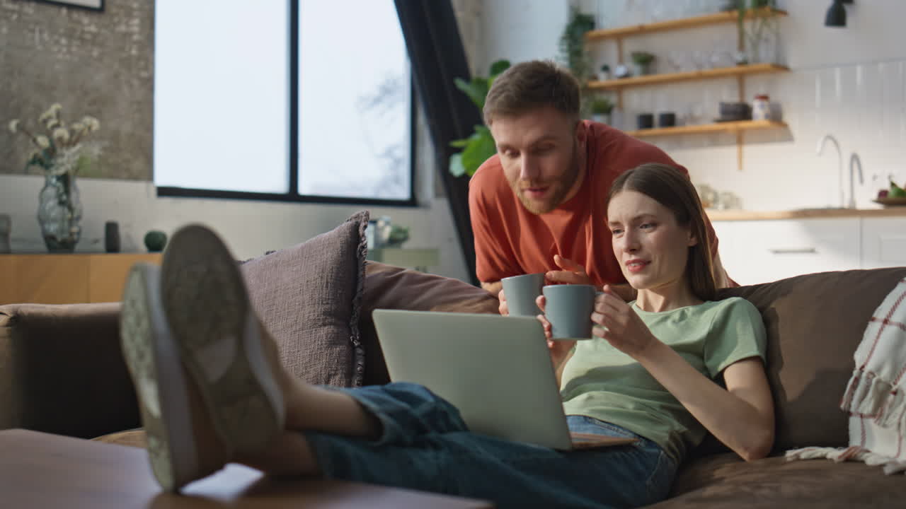 Woman working laptop lying comfortable at home couch. Husband bringing coffee