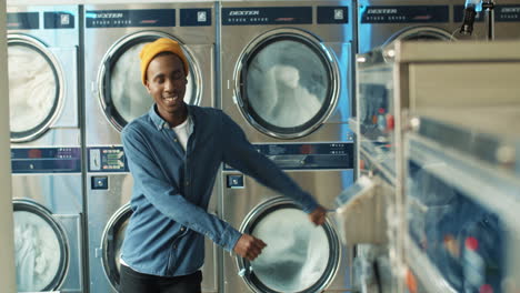 young stylish african american cheerful man having fun and dancing in laundry service room while machines washing on background. handsome guy listening to music while machines working in washhouse.