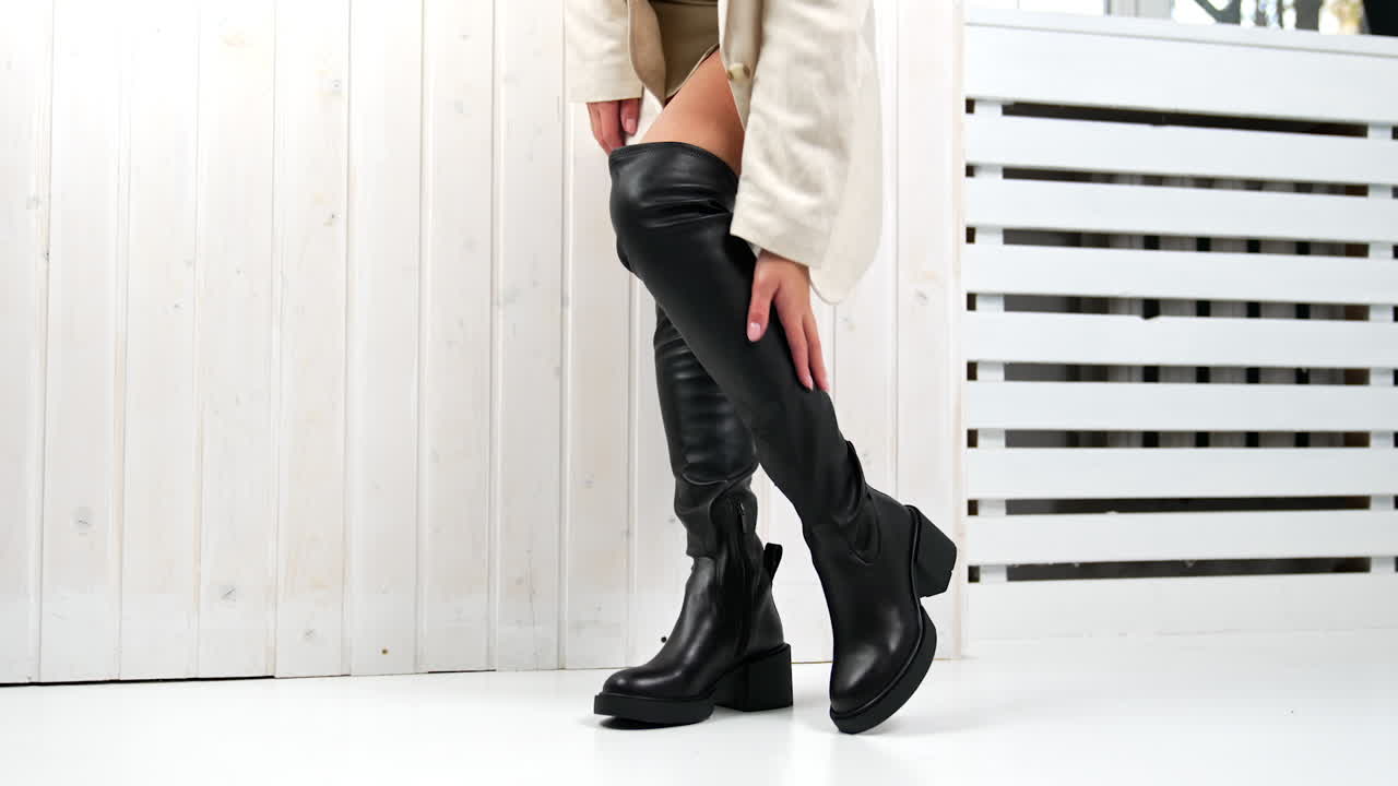 Elegant very high black boots demonstration in studio. Model in white mini skirt touching her footwear standing at the white wall.