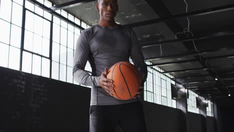 hombre afroamericano de pie en un edificio vacío jugando con una pelota de baloncesto mirando a la cámara