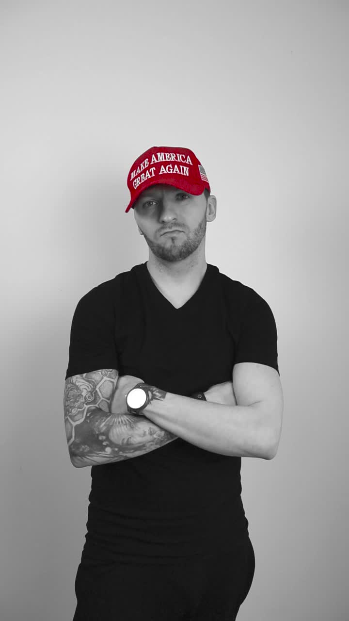 Grayscale footage of a man with tattoos wearing a red colored MAGA Make America Great Again Trump hat crossing his arms, watching directly forward and nodding with approval, showing he is a republican