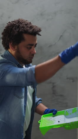 curly haired african-american male painter applies paint from tray on wall in living room close view. black man decorator changes color in new apartment