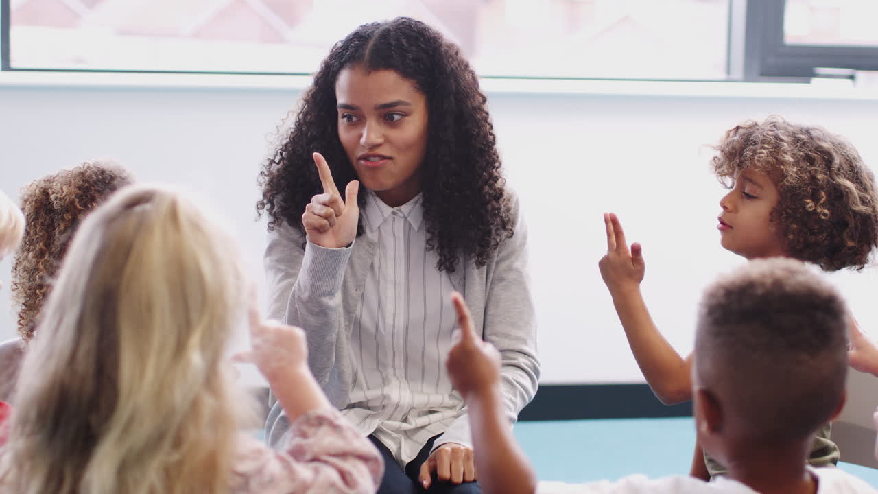 Infant school children sitting in class raising hands, learning to count with their female teacher