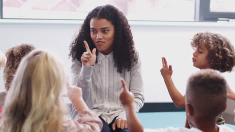 infant school children sitting in class raising hands, learning to count with their female teacher