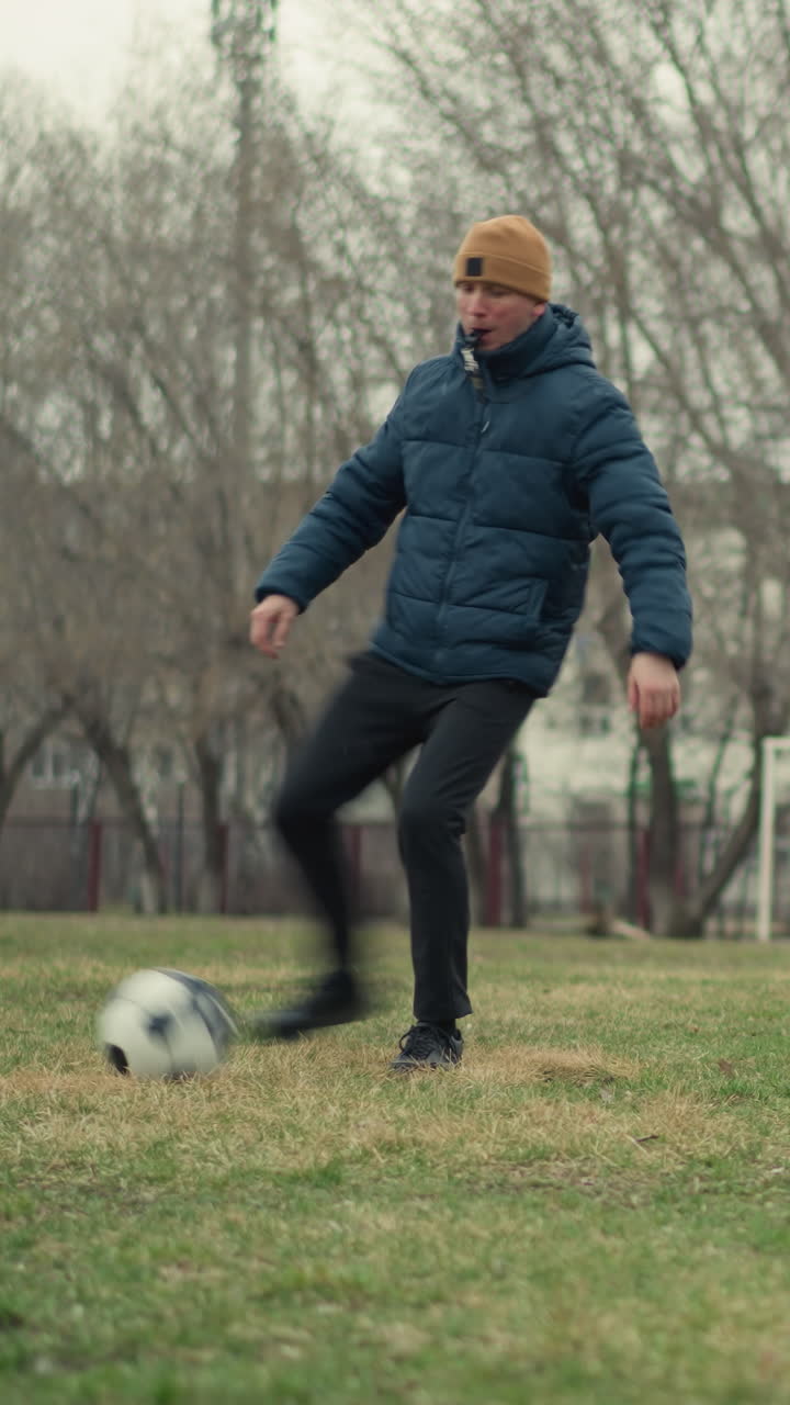 A soccer ball is passed to a coach, who has a whistle in his mouth, the coach skillfully plays with the ball while pointing out something, with trees and a goal post in the field