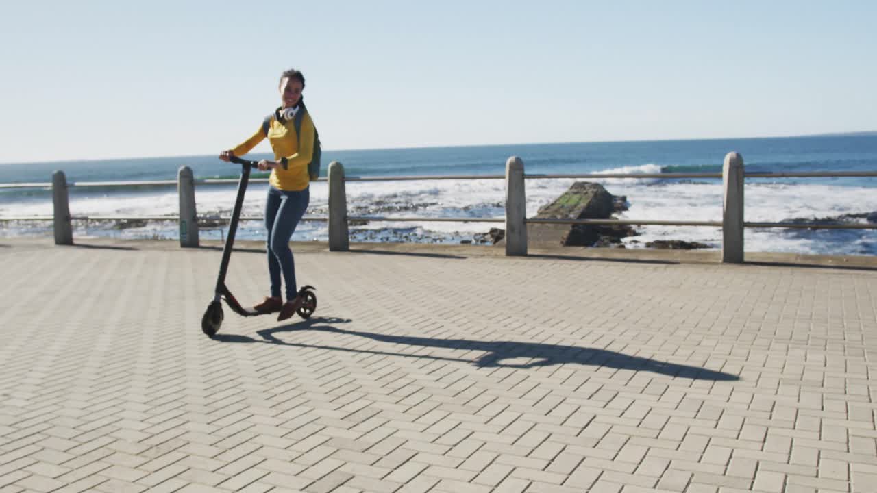 African american woman wearing headphones and backpack riding scooter on promenade by the sea