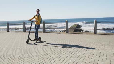 African-american-woman-wearing-headphones-and-backpack-riding-scooter-on-promenade-by-the-sea