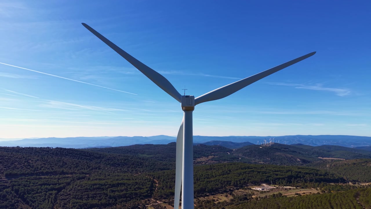 Vista posteriore ravvicinata di una turbina eolica a tre pale contro un cielo blu, circondata da colline boscose e un parco eolico distante all'orizzonte.