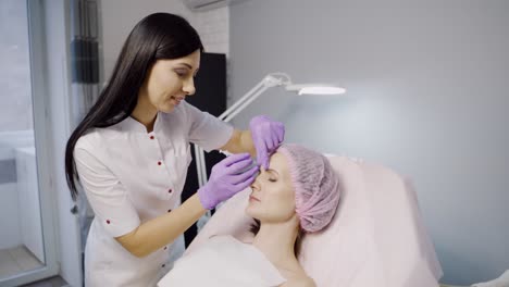 a pretty cosmetologist with a dark hair color makes botox for a woman who lays on the couch in a beauty salon. botulinum toxin