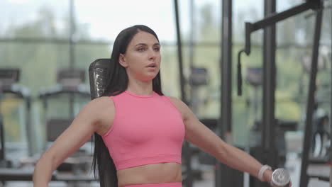 Una-Mujer-Morena-Con-Un-Traje-Rosa-Levanta-Pesas-A-Los-Lados-Mientras-Entrena-Sus-Hombros-En-El-Gimnasio.-Ejercicio-En-Banco-De-Hombros-Y-Brazos-Sentado.