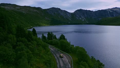 Toma-Aérea-Siguiendo-Un-Camión-Dorado-A-Través-Del-Paisaje-De-árboles-Verdes-En-Lofoten,-Norte-De-Noruega-Hacia-Nusfjord-Con-El-Camino-Serpenteando-A-Través-De-Lagos-Con-Montañas-Dramáticas-En-El-Fondo