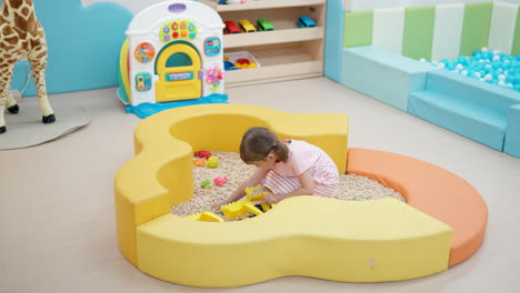 child girl in playroom loading sand in yellow toy excavator bucket or scoop - wide angle