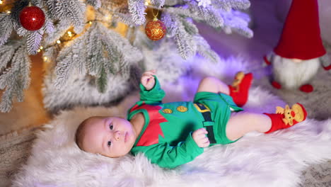 sweet little boy in green elf costume lies on white plaid under beautifully decorated christmas tree. cute child tosses his feet actively and looks into camera.
