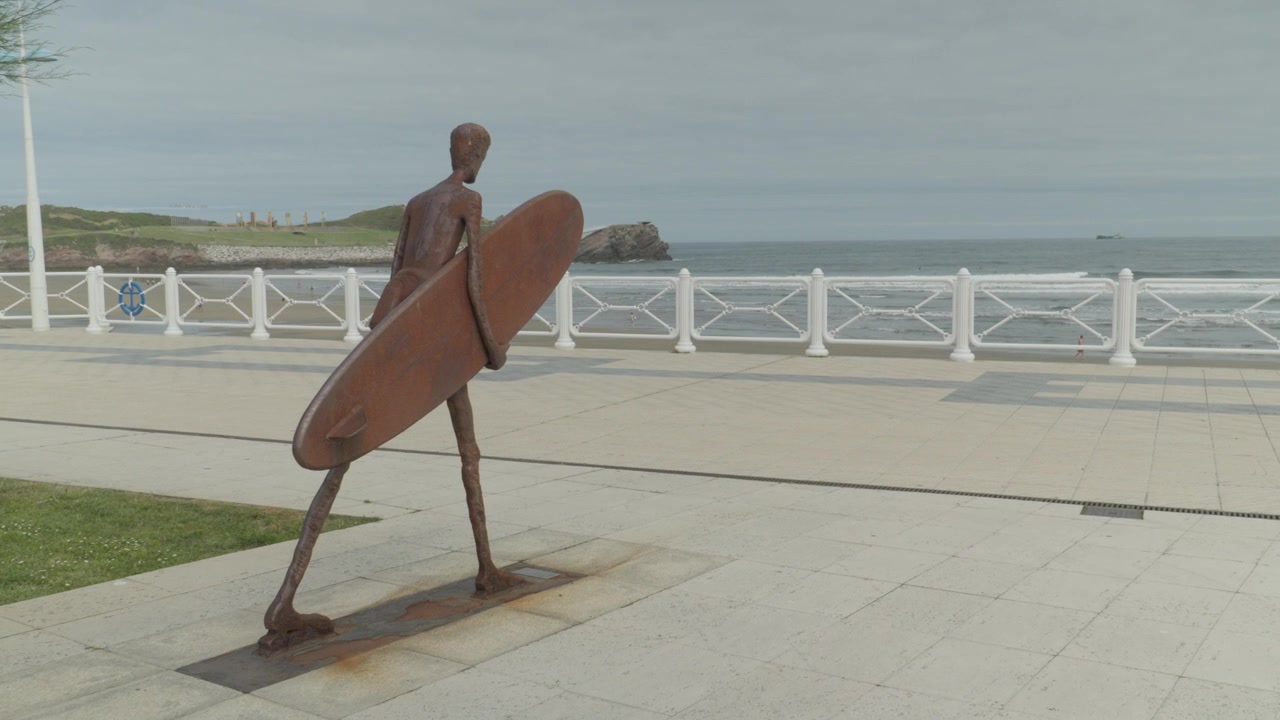 Carrellata del famoso monumento in bronzo del surfista sul lungomare di Salinas, Asturie, Spagna. Mostra la scultura stilizzata, la piazza e l'edificio della scuola di surf locale sotto un cielo luminoso