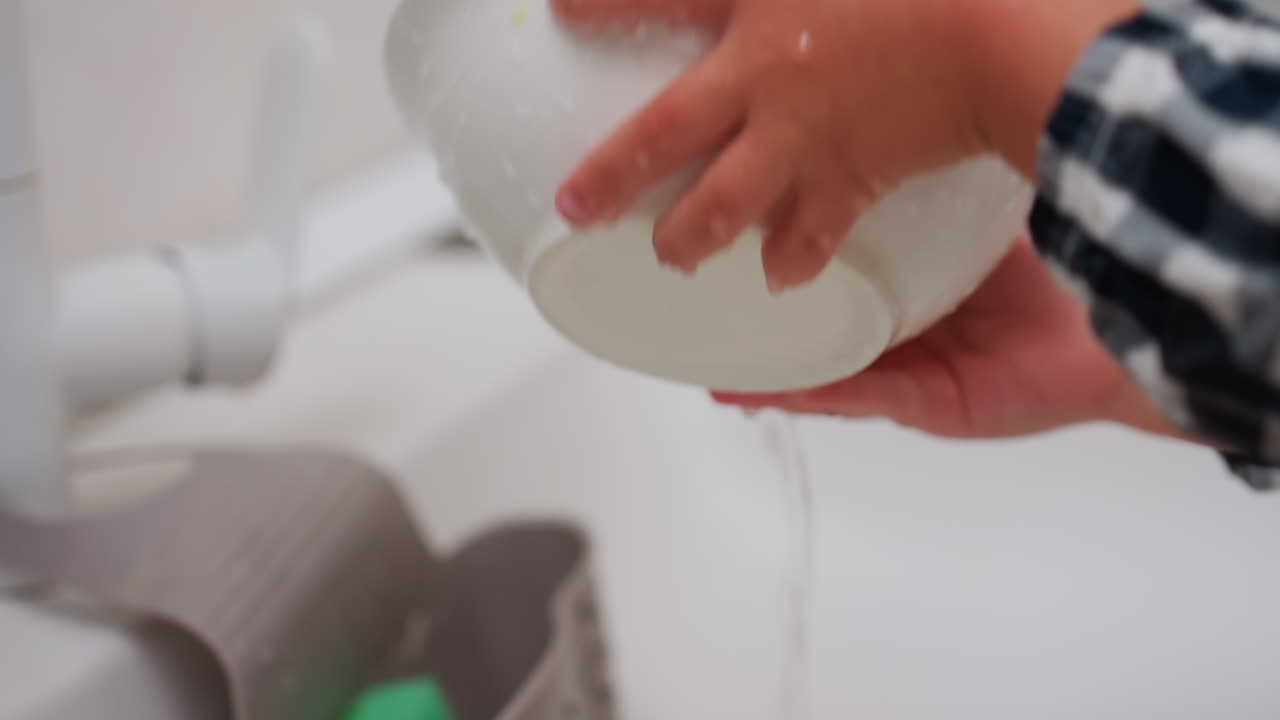 Close up of toddler rinsing plate under flowing water at sink turning stream into basin with small hands holding dish carefully in checkered sleeve showing innocence learning responsibility