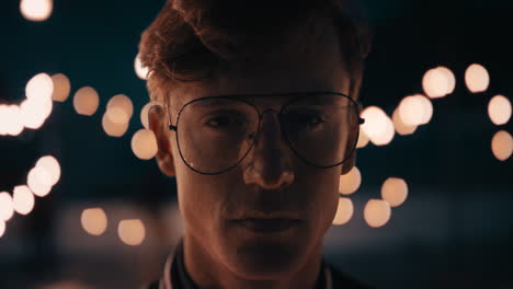 retrato joven pelirrojo con gafas en la azotea por la noche mirando una expresión seria con hermosas luces de fiesta en el fondo del concepto de juventud urbana