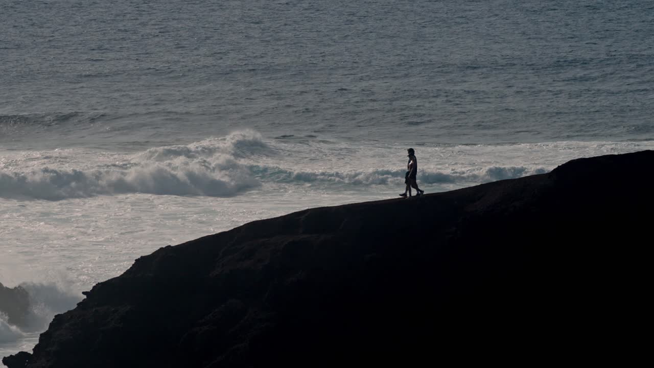 A young couple walks hand in hand along a rugged volcanic cliff overlooking the Atlantic Ocean in Lanzarote, Canary Islands, Spain.