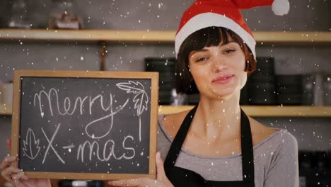 video composition with falling snow over cafe shop owner in santa hat holding board with greetings