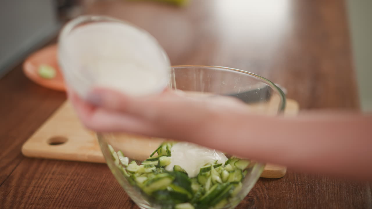 close up of fair skinned person holding transparent bowl while pouring salad cream into freshly sliced cucumber inside clear glass container placed on wooden kitchen counter with cutting board nearby