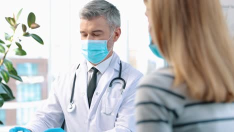 Close-Up-Portrait-Of-Handsome-Middle-Aged-Experienced-Male-Doctor-Immunologist-Looking-At-The-Patient-And-Speaking-In-Cabinet-In-Hospital,-While-Showing-Covid-Vaccines-To-The-Woman